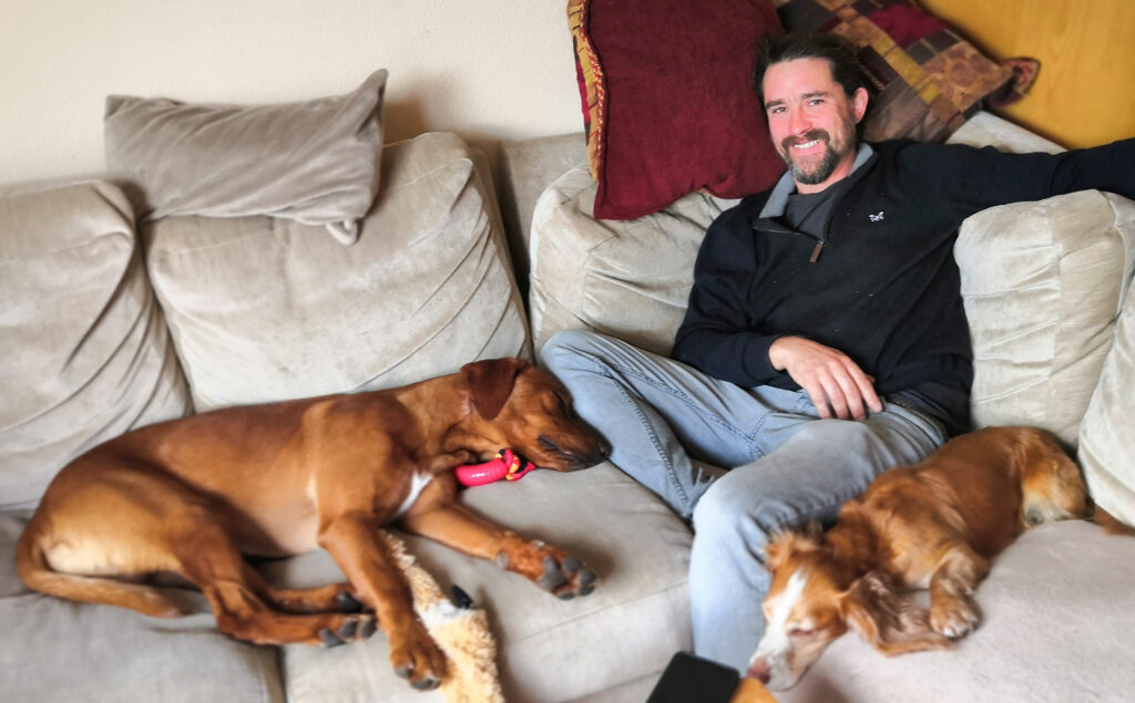 a man called Andrew Parsons sitting on a cream sofa with a fox red labrador dog and a red coloured working cocker spaniel
